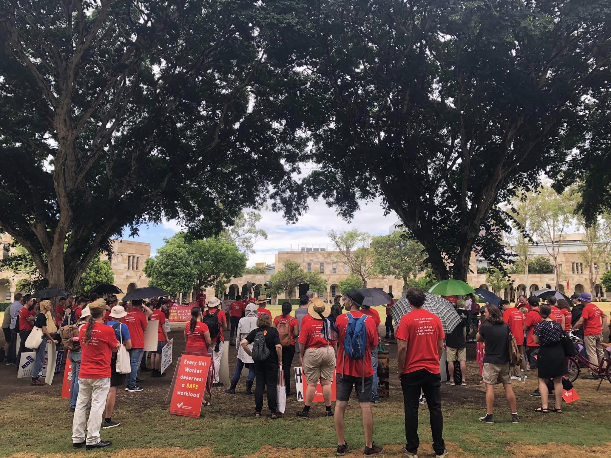 A crowd of protestors in red NTEU shirts standing and facing away from the camera. They are under trees in UQ's Great Court.